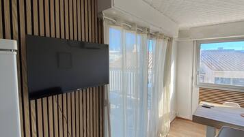 A modern living room with a flat-screen TV mounted on a wooden panel wall, a dining area with a table and chairs, and a large window with curtains.