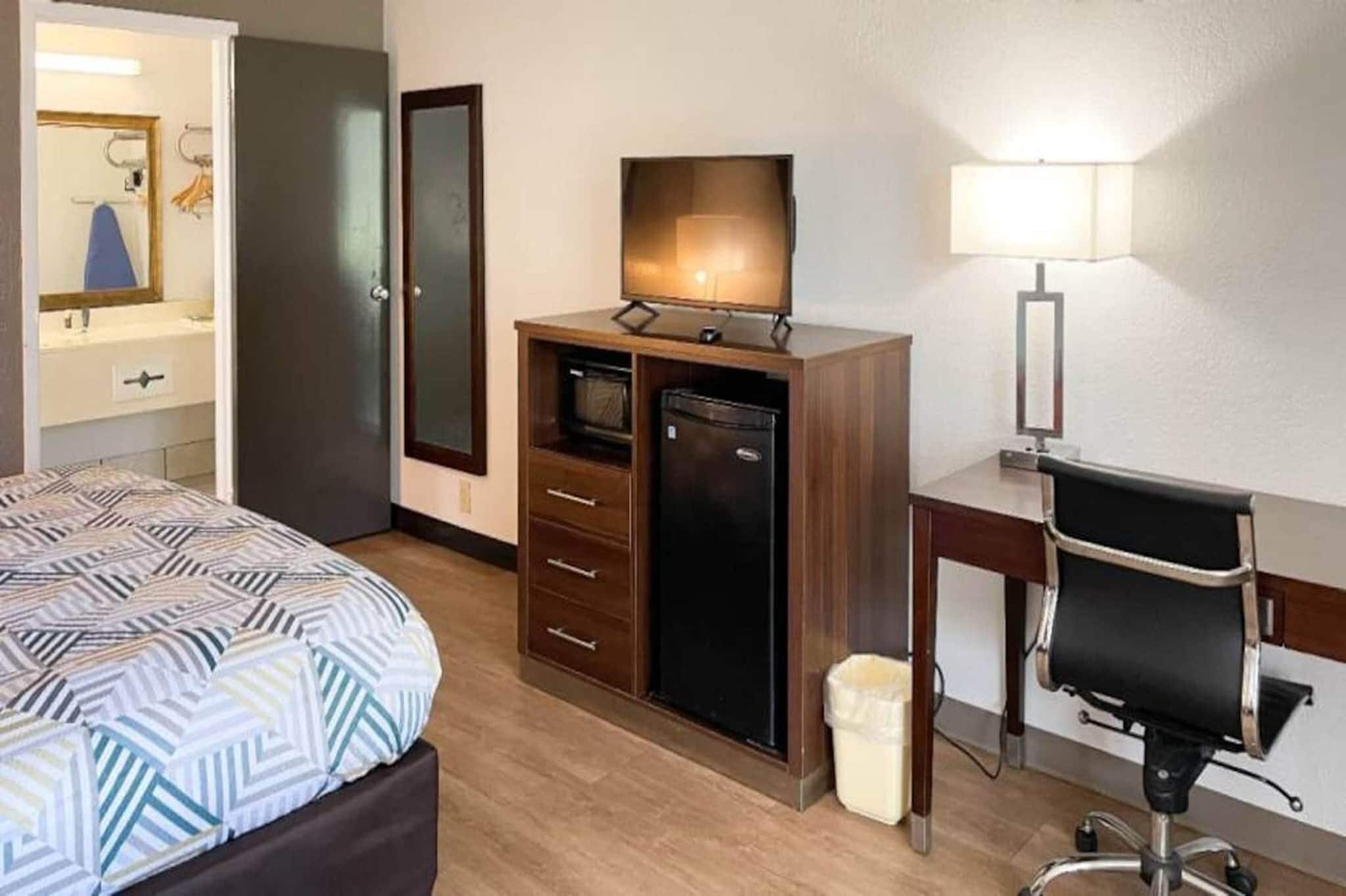 Quarto de hotel com cama, escrivaninha, cadeira, televisão, luminária e armário.