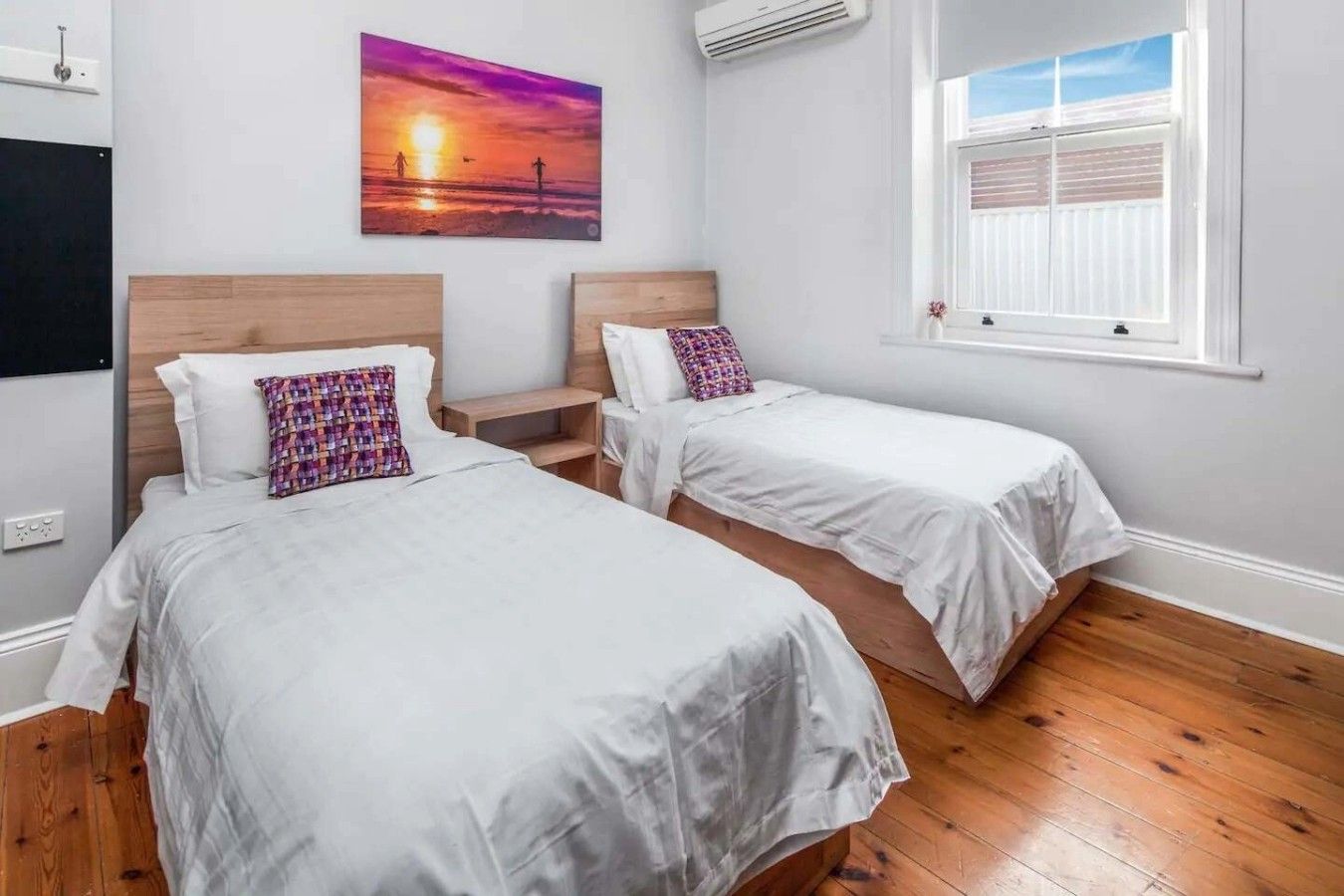 Quarto com duas camas, um quadro de um pôr do sol, ar condicionado e janela com persianas.