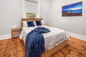 Een slaapkamer met een houten vloer, een bed met wit beddengoed en blauwe kussens, een nachtkastje met een lamp, en een schilderij aan de muur.