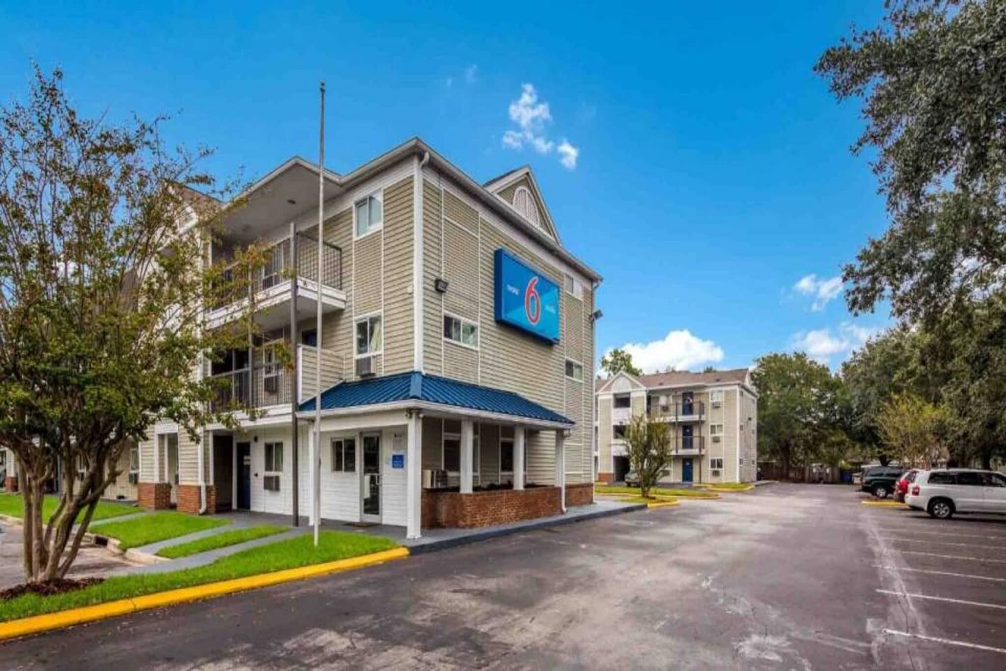 A multi-story hotel with a blue awning and a '6' sign, featuring a parking lot and greenery.
