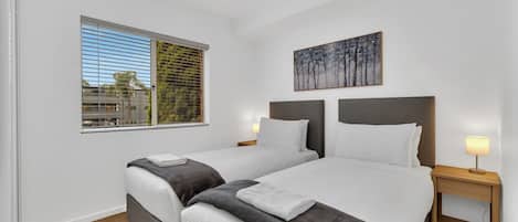 Une chambre d’hôtel avec deux lits, une fenêtre avec des stores, un tableau au mur et une table de chevet avec une lampe.