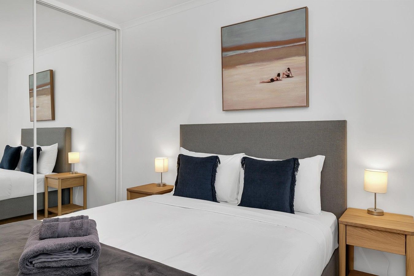 A hotel room with a bed, two bedside tables, a nightstand, a mirror, and a painting of a beach scene.