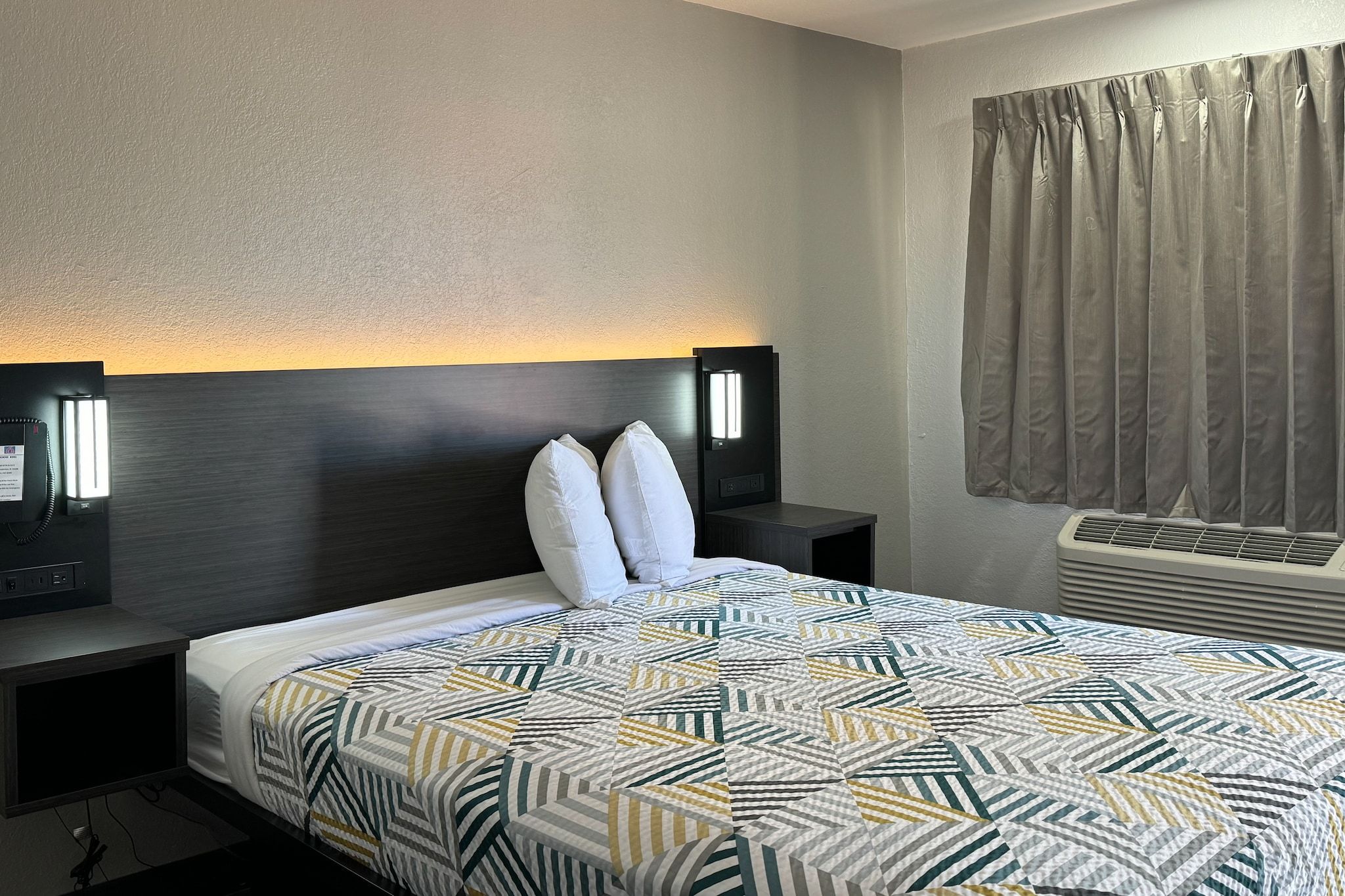 A hotel room with a bed, two bedside tables with lamps, a window with curtains, and an air conditioning unit.