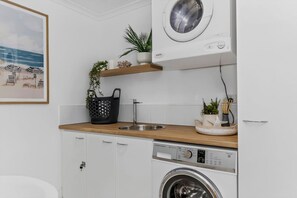 A modern kitchen with a built-in washing machine, a sink, and a countertop with a plant and a basket. - Colley Reserve Retreat (Glenelg)