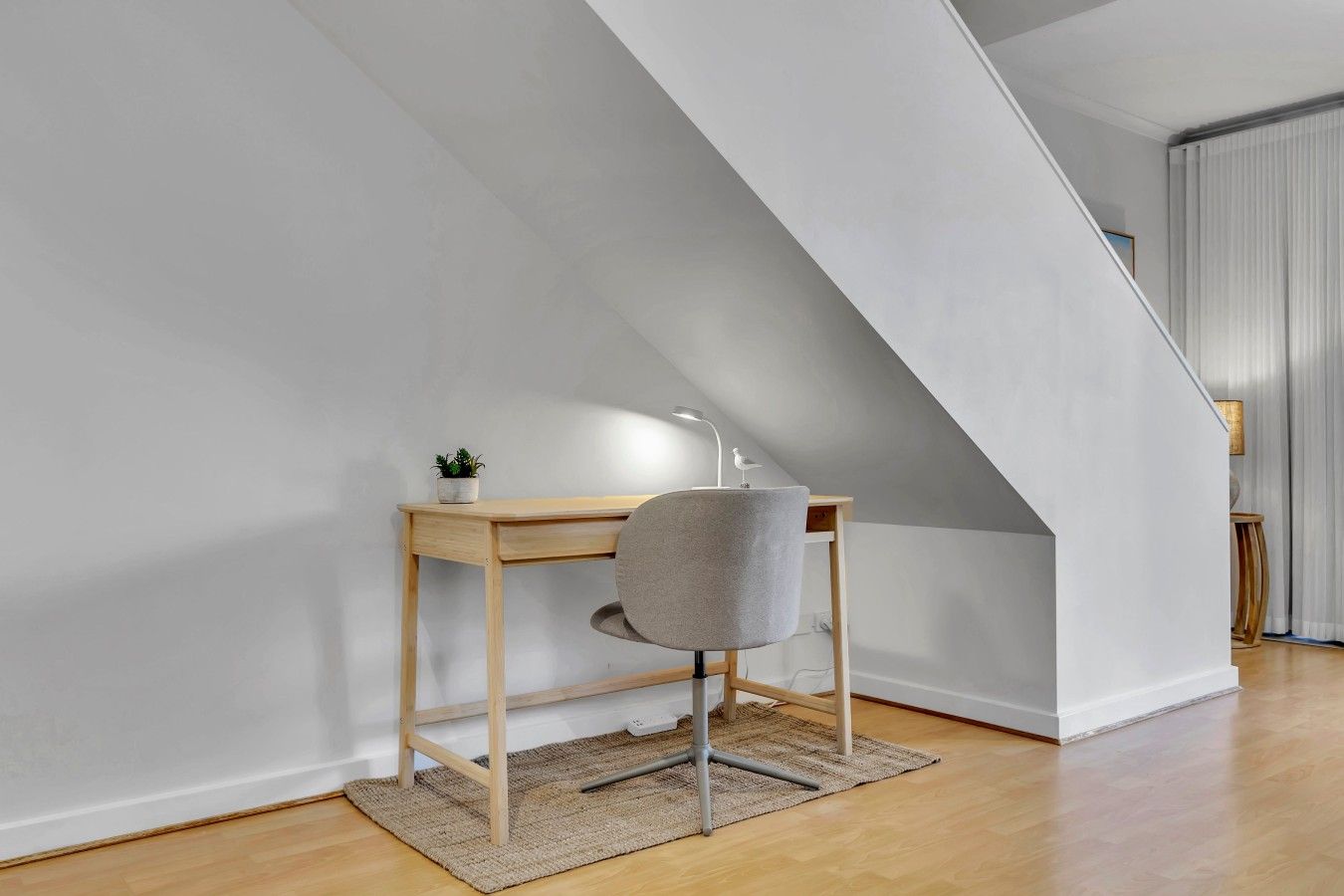 A modern loft bedroom with a sloped ceiling, a wooden desk, a grey chair, and a small potted plant.