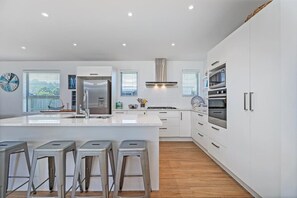 A modern kitchen with a central island, bar stools, stainless steel appliances, and white cabinetry. - Within Reach - Coastal Escape - Cooks Beach (Cooks Beach)