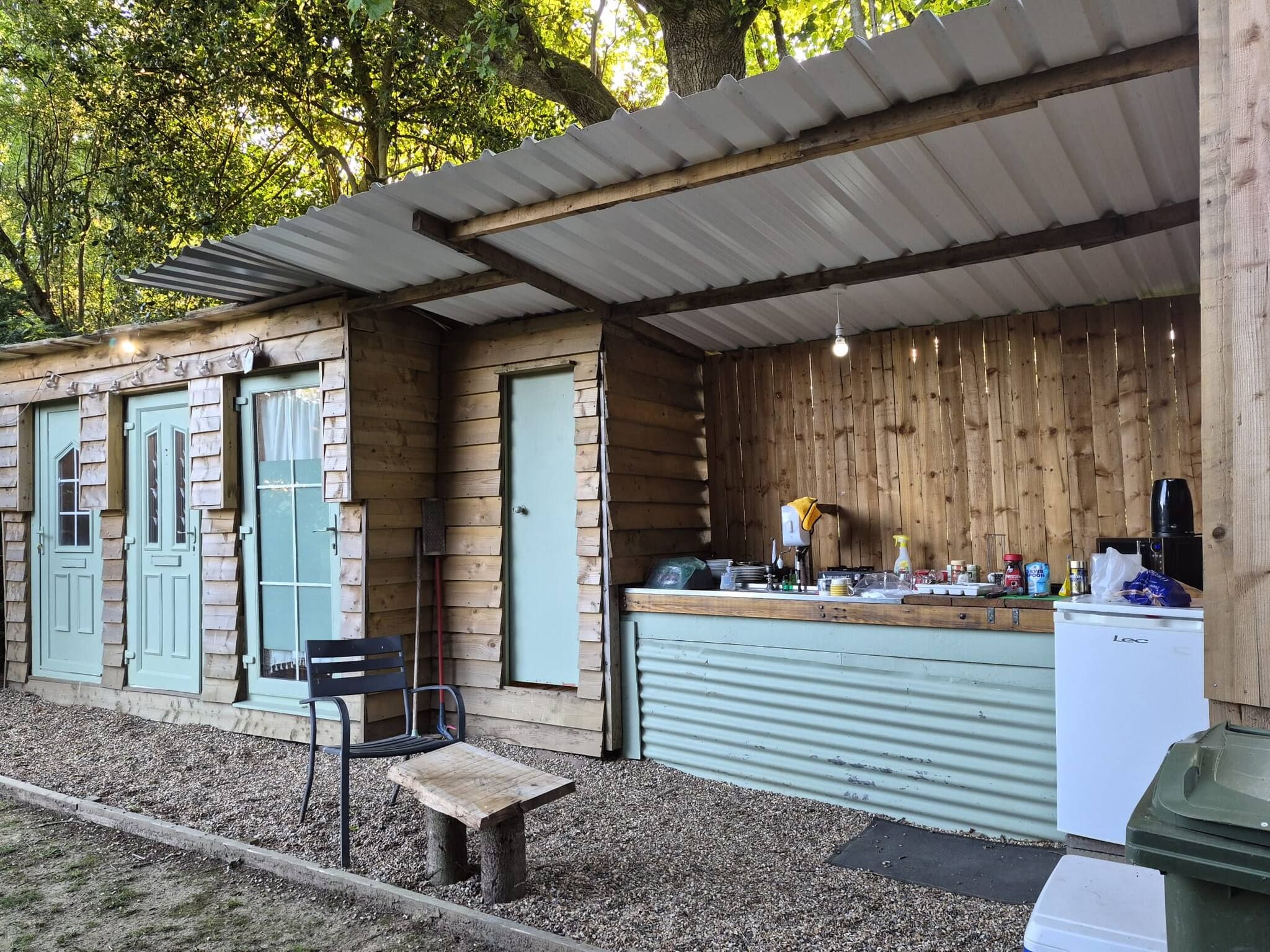 Une structure rustique en bois comprenant un espace cuisine, un banc et une poubelle.