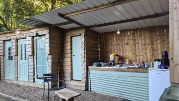 Une structure rustique en bois comprenant un espace cuisine, un banc et une poubelle.