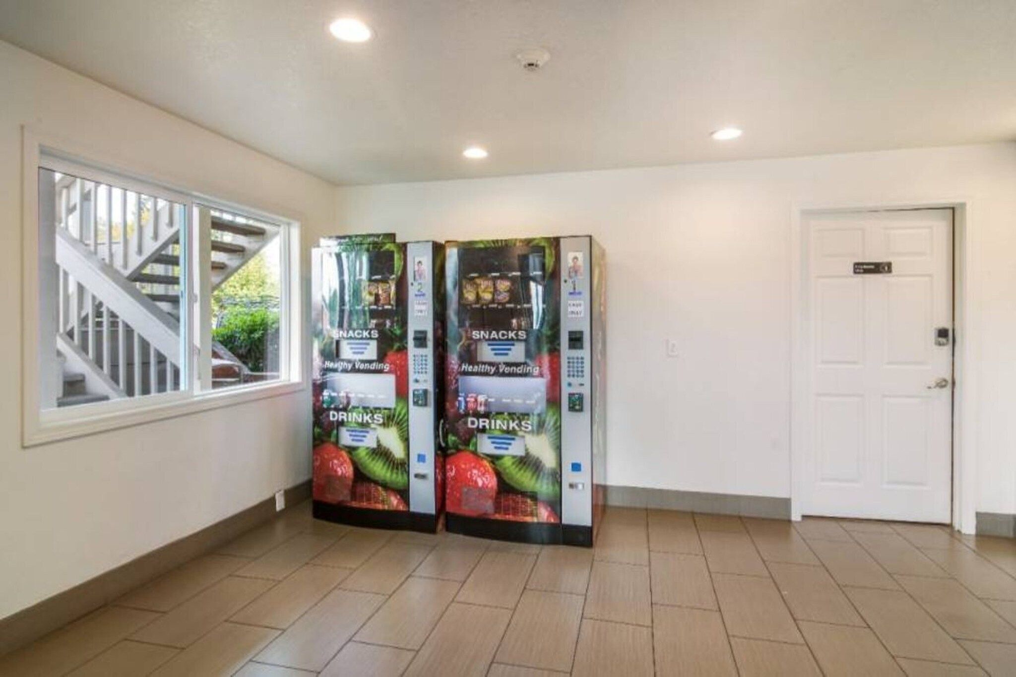 A hallway with two vending machines labeled 'Healthy Vending' for snacks and drinks, a staircase leading to an upper level, and a white door.
