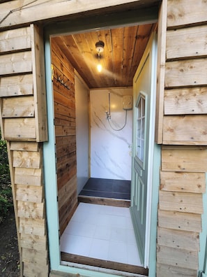 A wooden cabin with a small bathroom featuring a shower, a tiled floor, and a wooden door. - The Bird Nest at The Secret Garden (Biddenden)