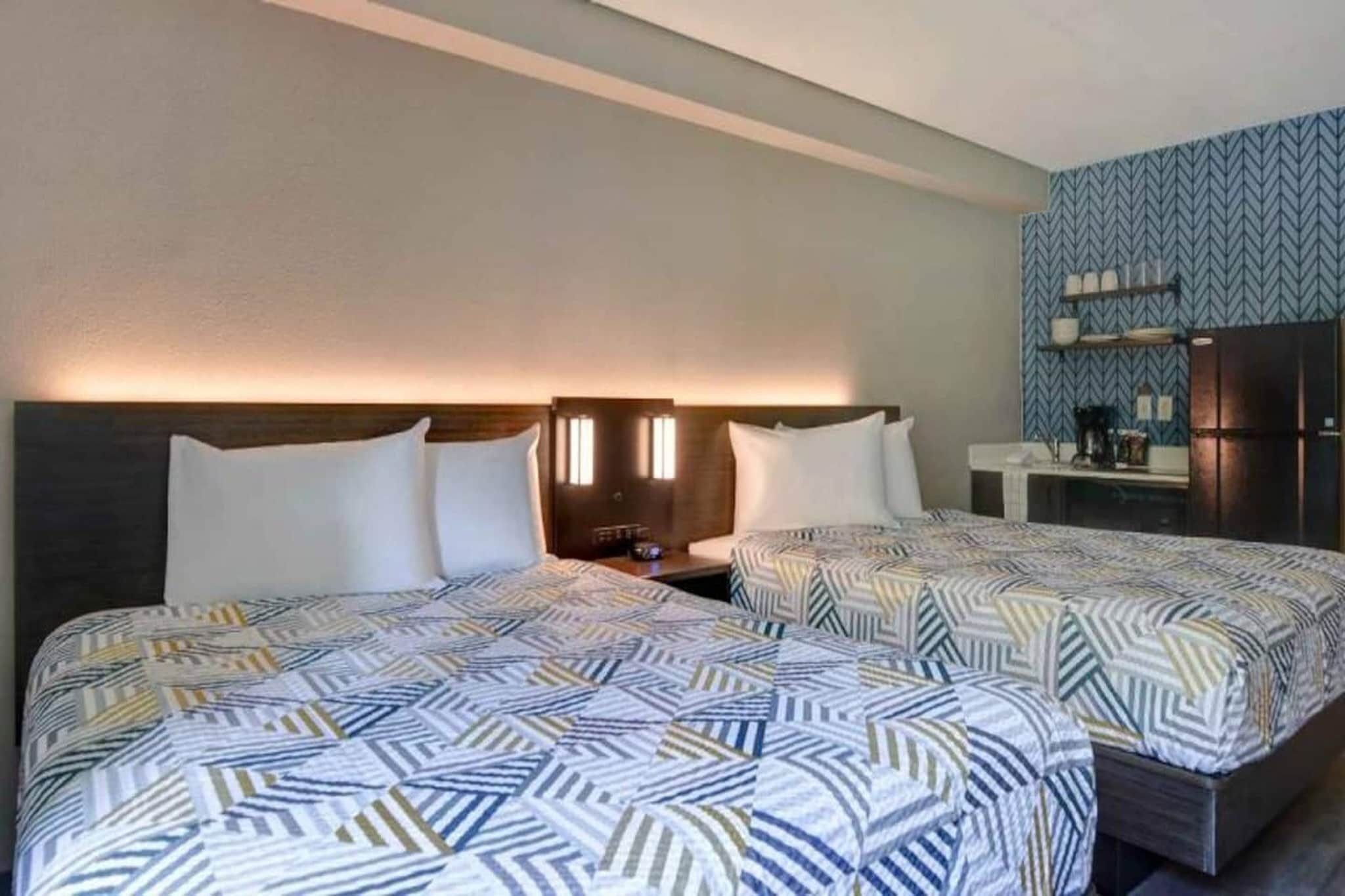 A hotel room with two beds, a nightstand, a small table, and a refrigerator.