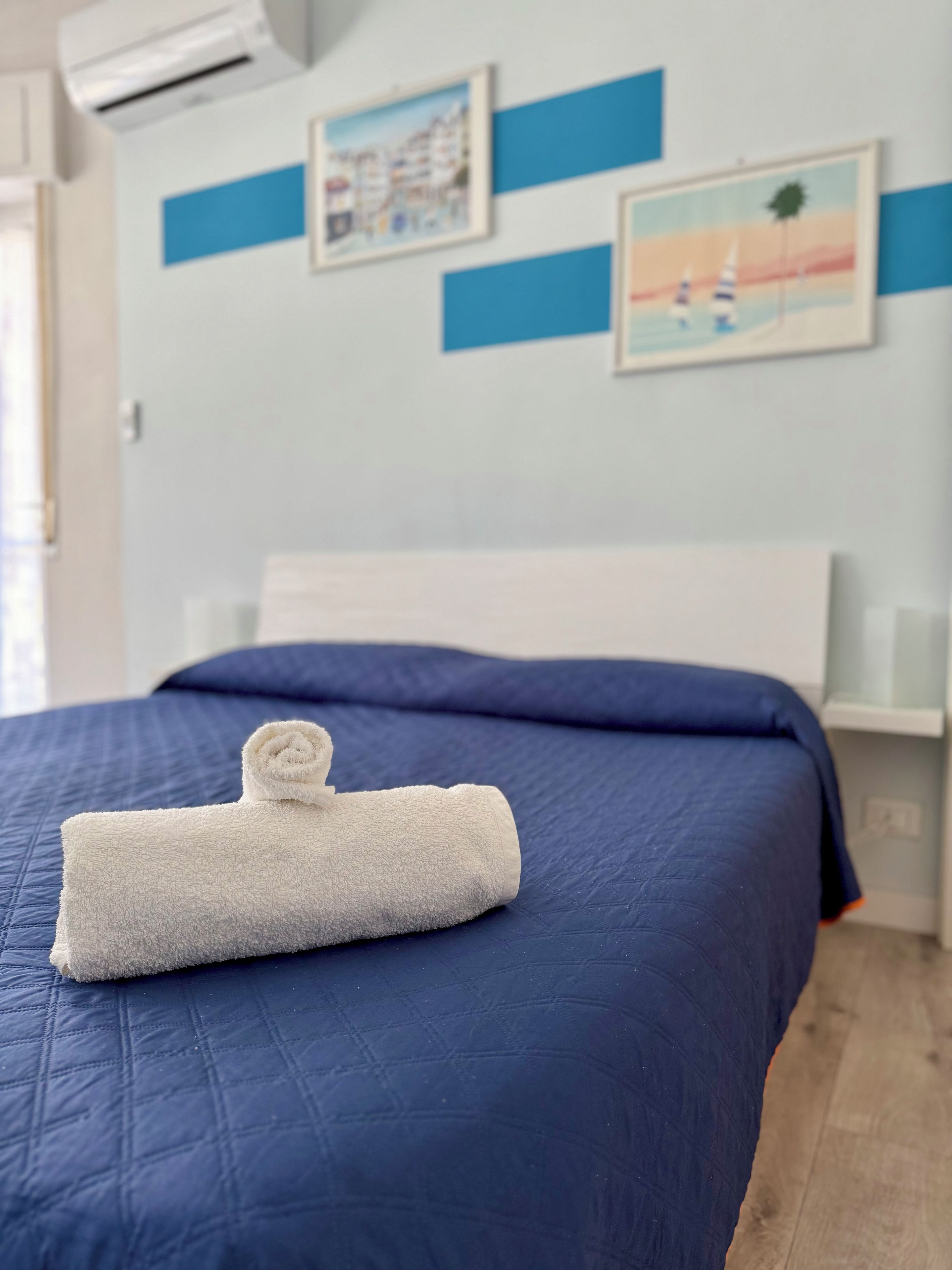 Siististi pedattu sänky, jossa on rullattu valkoinen pyyhe, sininen päiväpeite ja valkoinen pääty.