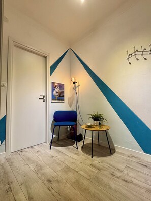 A room with a door, a blue chair, a small table with a plant, and a wall with a geometric design. - CasaFerruccio [Private Parking - Air Conditioning - Wi-Fi] (Sanremo)