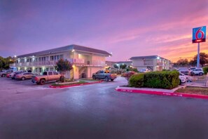 Un motel con estacionamiento, un cartel con el número 6 y un edificio de techo plano.