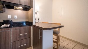 Cocina compacta con armarios de madera oscura, encimera negra y un pequeño comedor con una silla.