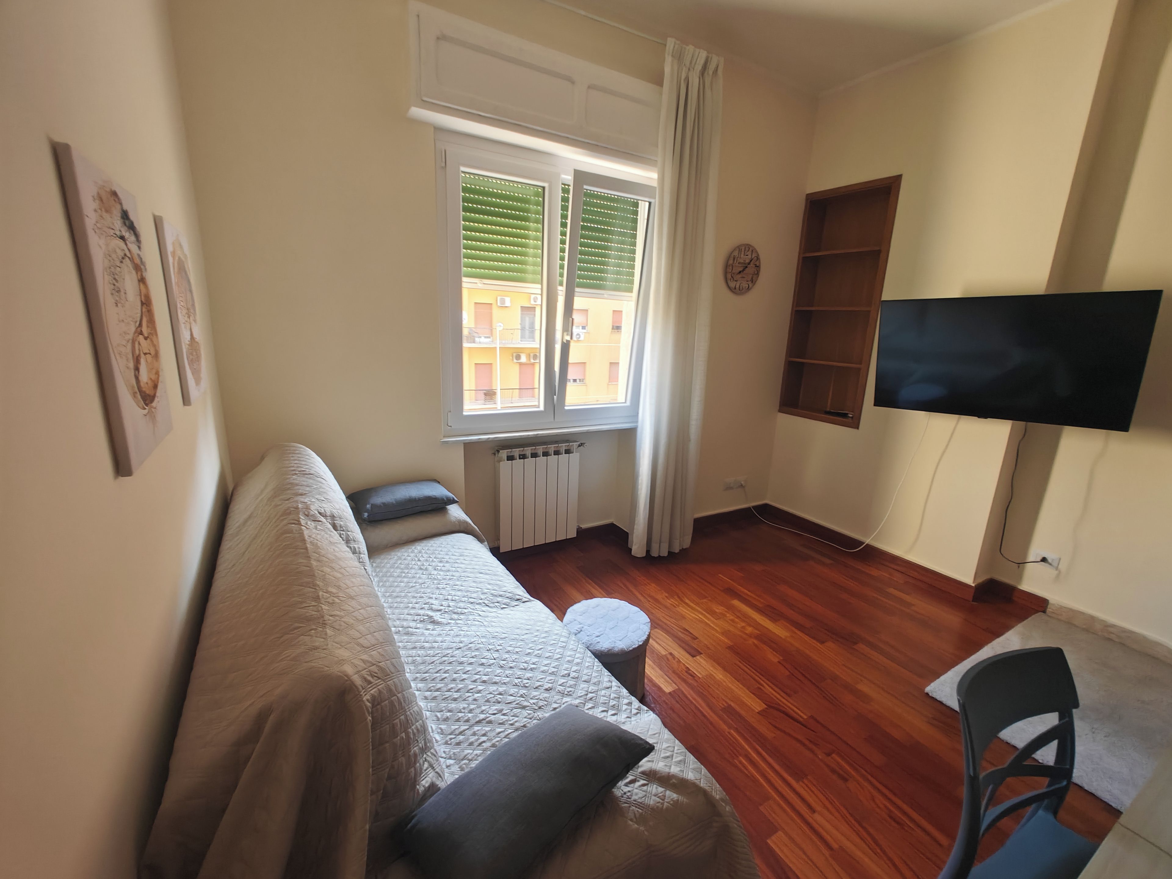 Ein möbliertes Wohnzimmer mit einer Couch, einem an der Wand montierten Fernseher, einem Fenster mit Jalousien und einer Heizung.
