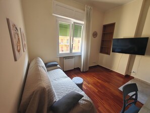 Un salon meublé comprenant un canapé, un téléviseur fixé au mur, une fenêtre avec des stores et un radiateur.