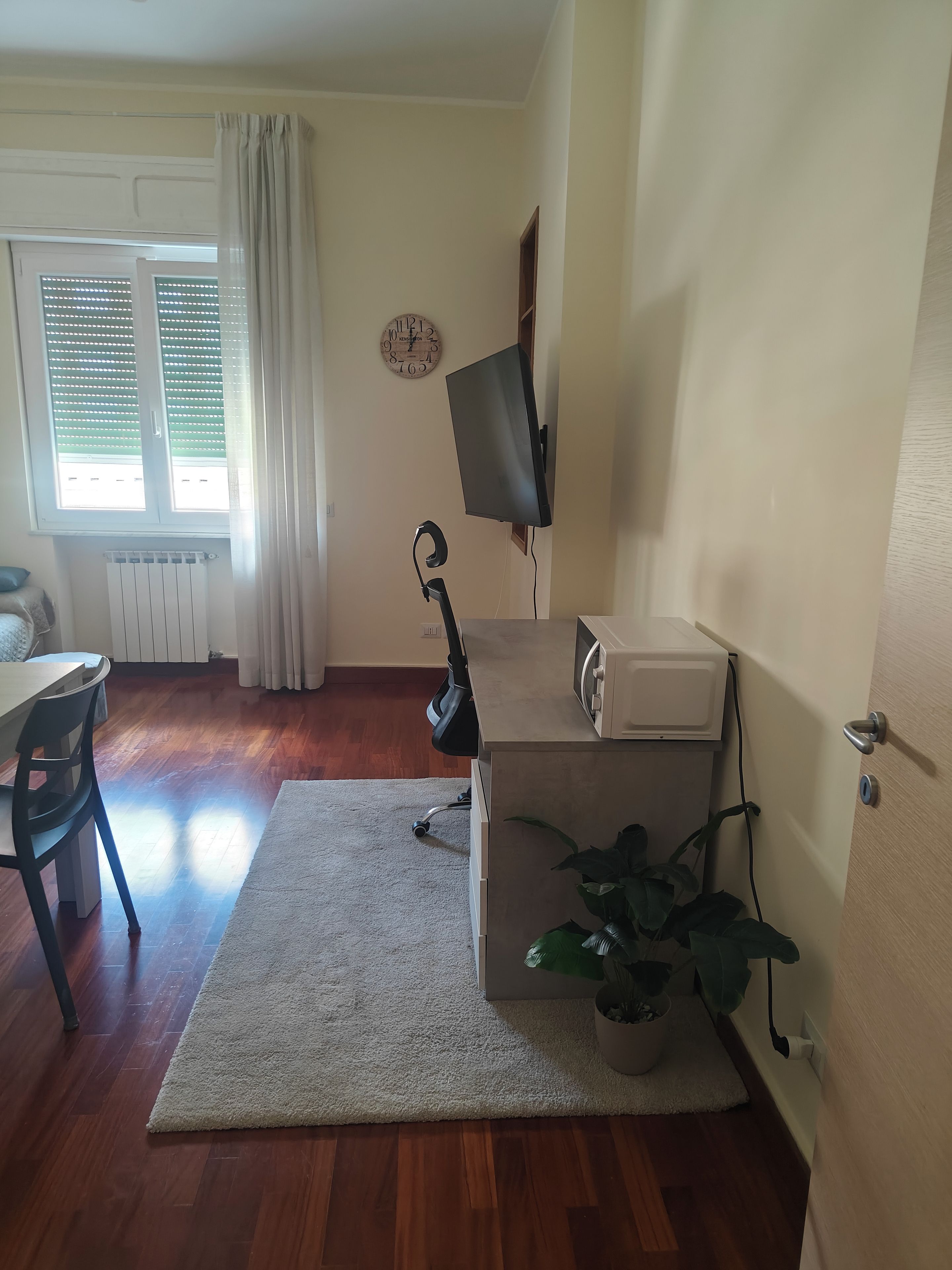 Ein Zimmer mit Schreibtisch, Stuhl, Heizkörper, Vorhängen, einer Uhr, einem Fernseher und einer Pflanze.