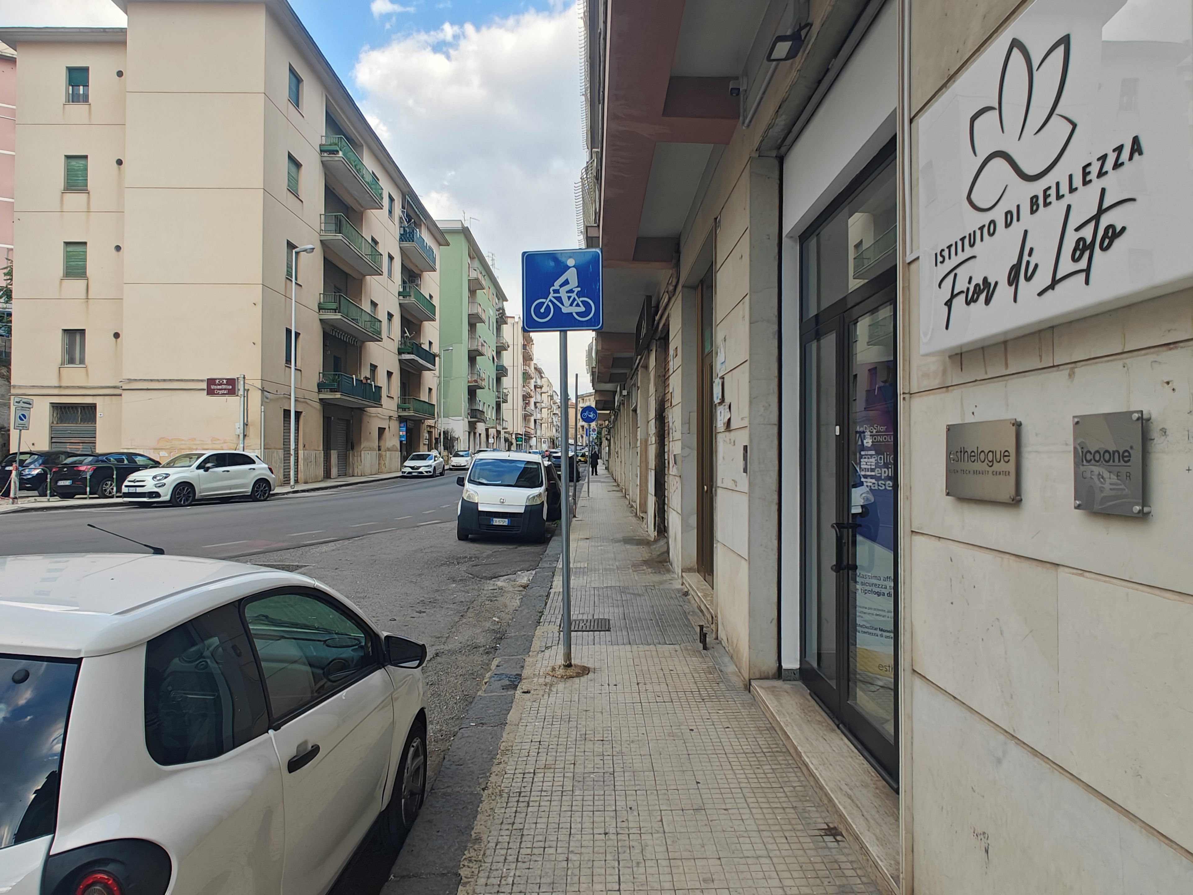 Une rue bordée de voitures garées, un trottoir et des immeubles avec des enseignes.