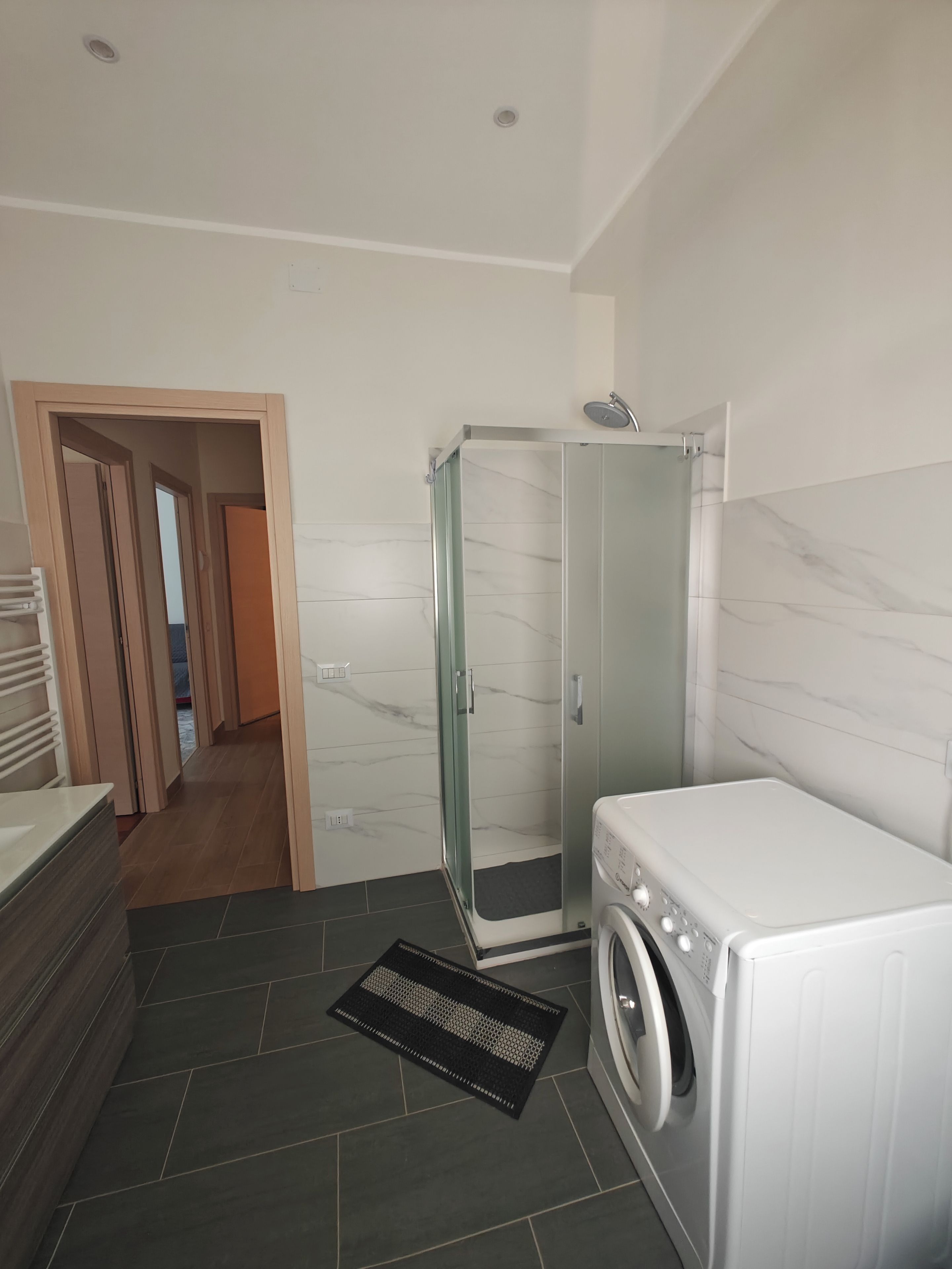Un baño moderno con cabina de ducha de vidrio, lavadora integrada y piso de baldosas oscuras.
