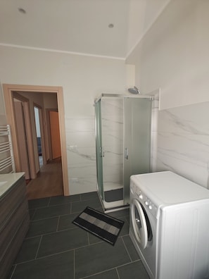 Un bany modern amb una cabina de dutxa de vidre, una rentadora integrada i un sòl de rajola fosca.