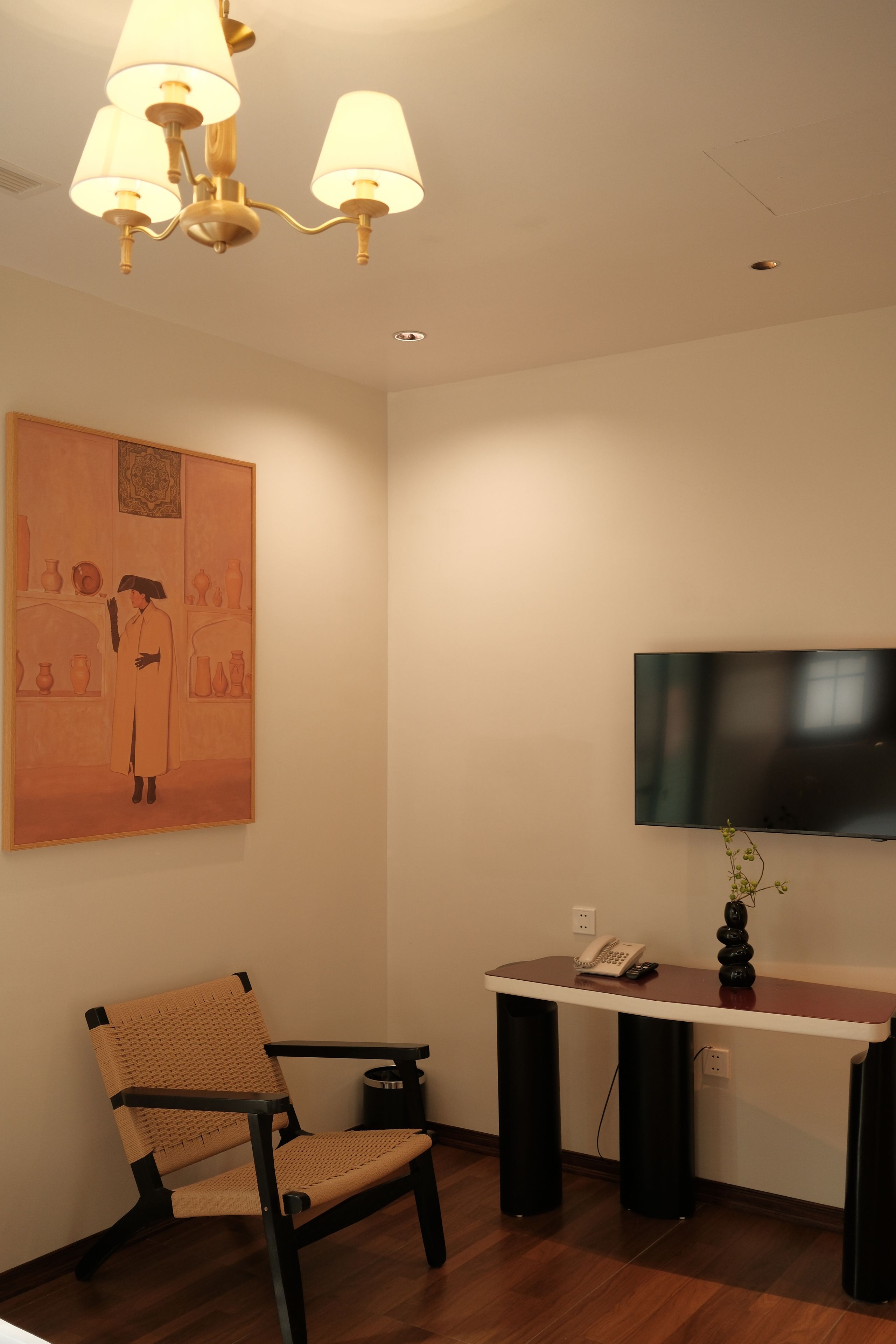 A room with a flat-screen TV mounted on the wall, a desk with a telephone, a woven chair, and a painting on the wall.