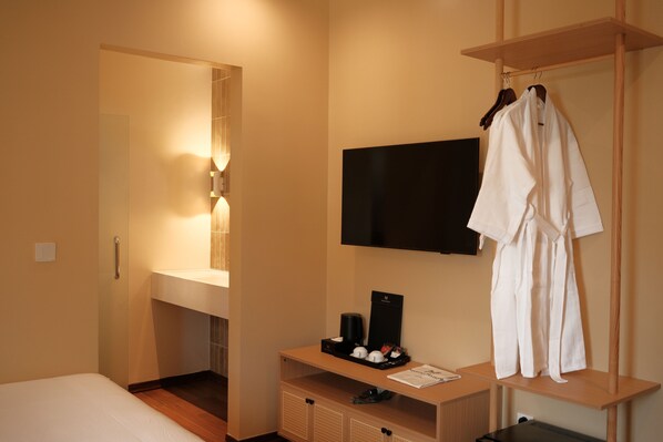Habitación de hotel con cama, tocador con espejo, televisor de pantalla plana montado en la pared, estante de madera con artículos de aseo y bata colgada en un perchero.