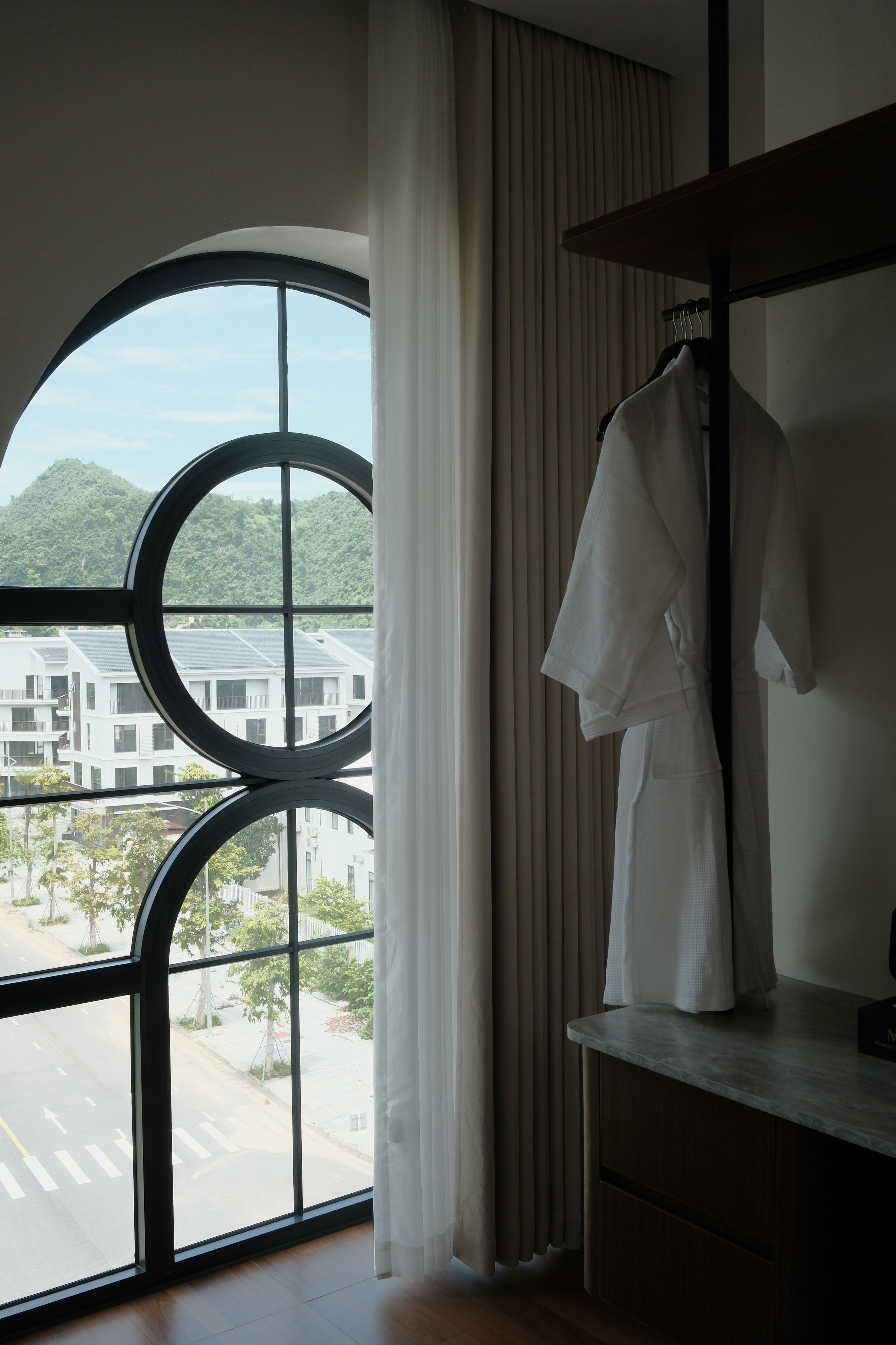 A hotel room with a large window, a robe hanging on a rack, and a view of a building and trees.