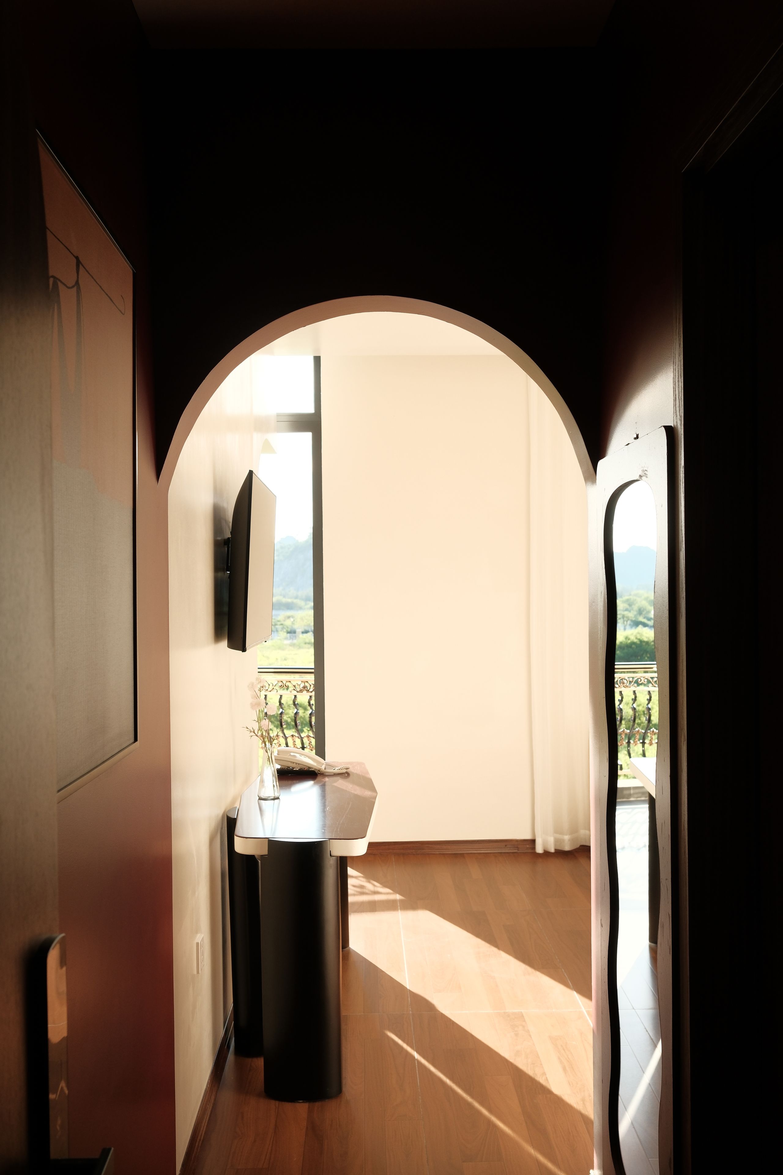 A modern interior with a wooden floor, a kitchen area with a sink and a vase of flowers, and a large arched doorway leading to a balcony with a view of greenery.