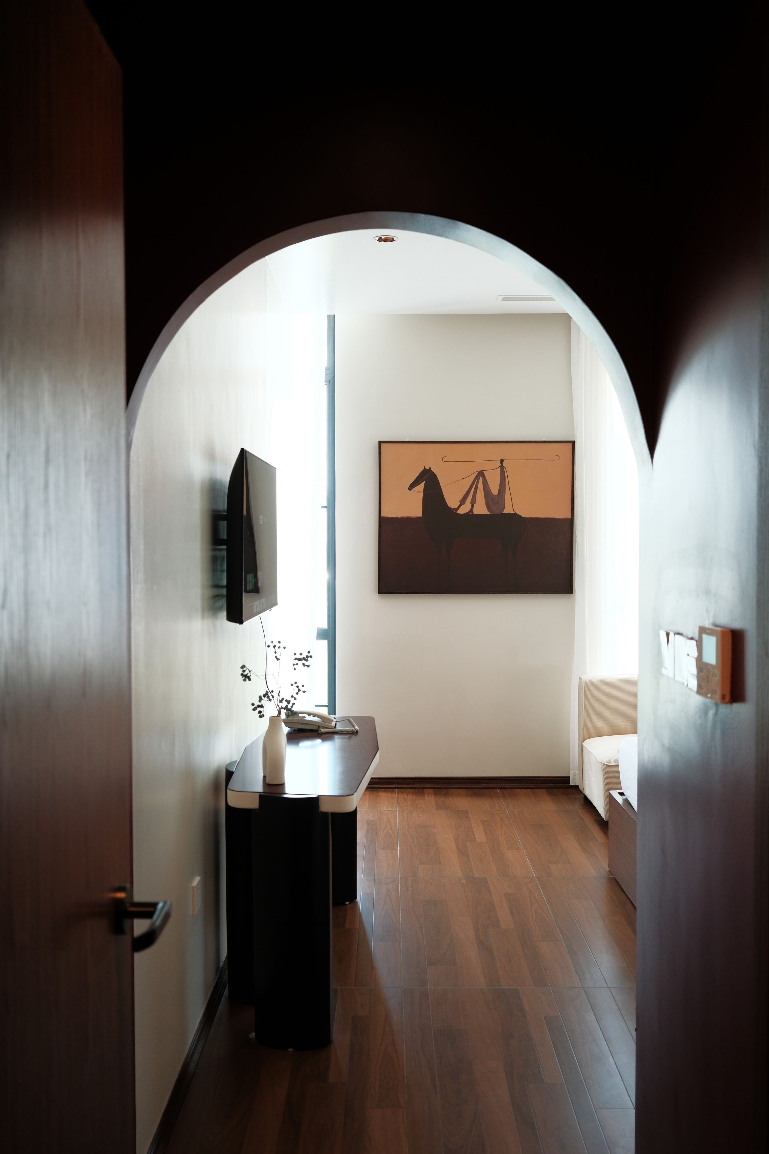 A hallway with a wooden floor, a small table with a vase and a phone, and a painting on the wall.