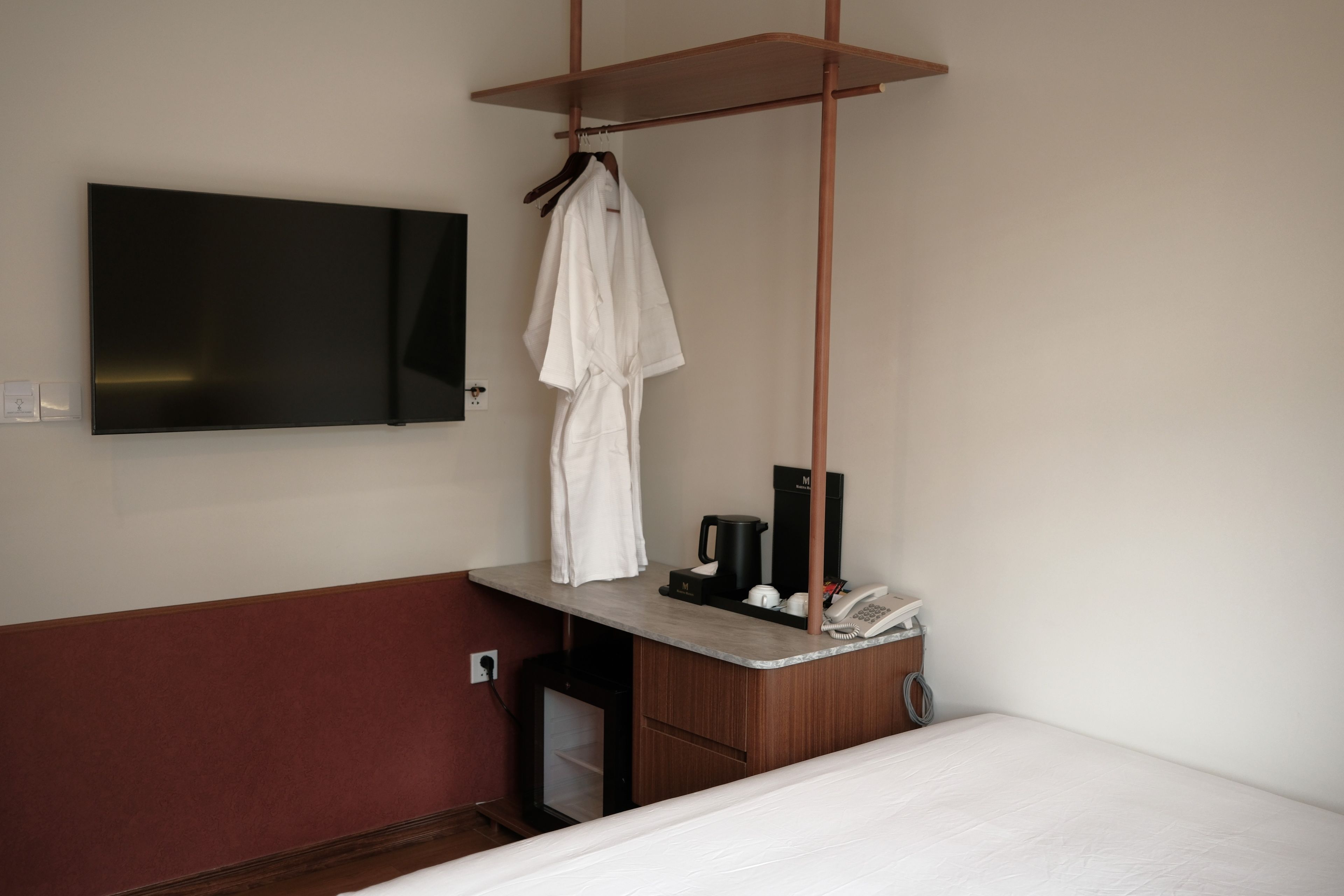 Habitación de hotel con cama, televisor montado en la pared, bata colgada en un perchero y una mesita con teléfono y cafetera.