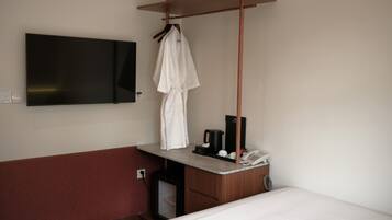 Habitación de hotel con cama, televisor montado en la pared, bata colgada en un perchero y una mesita con teléfono y cafetera.