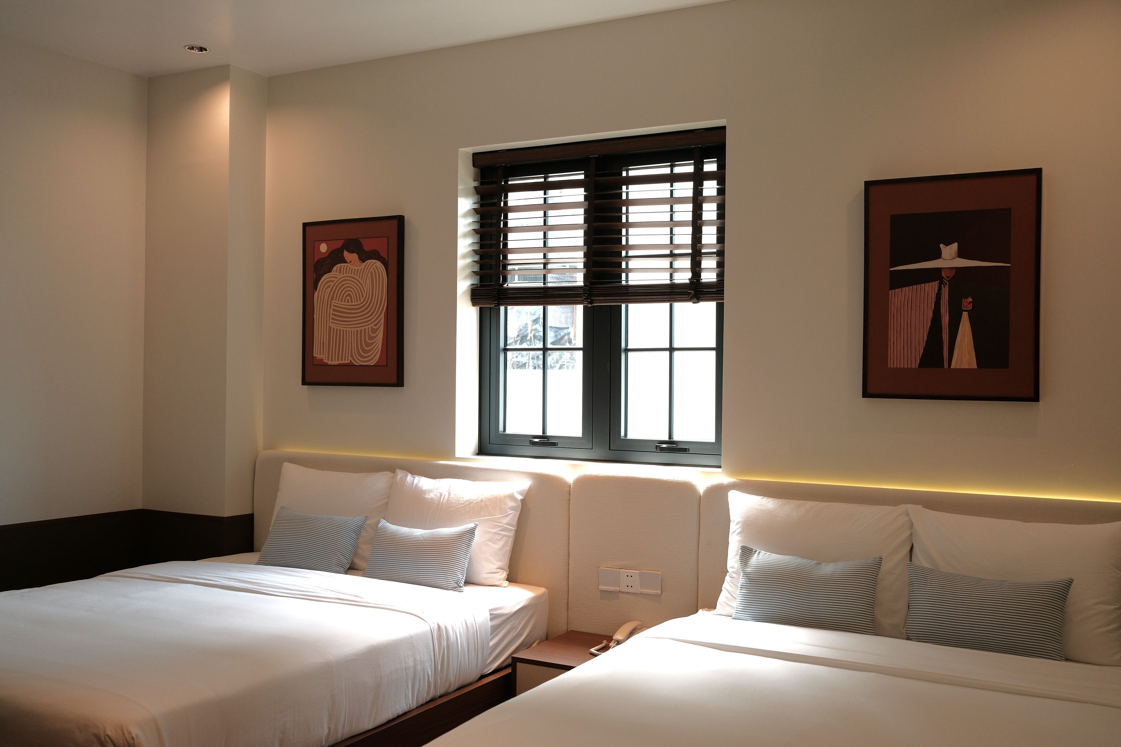 A hotel room with two beds, a window with blinds, and artwork on the walls.