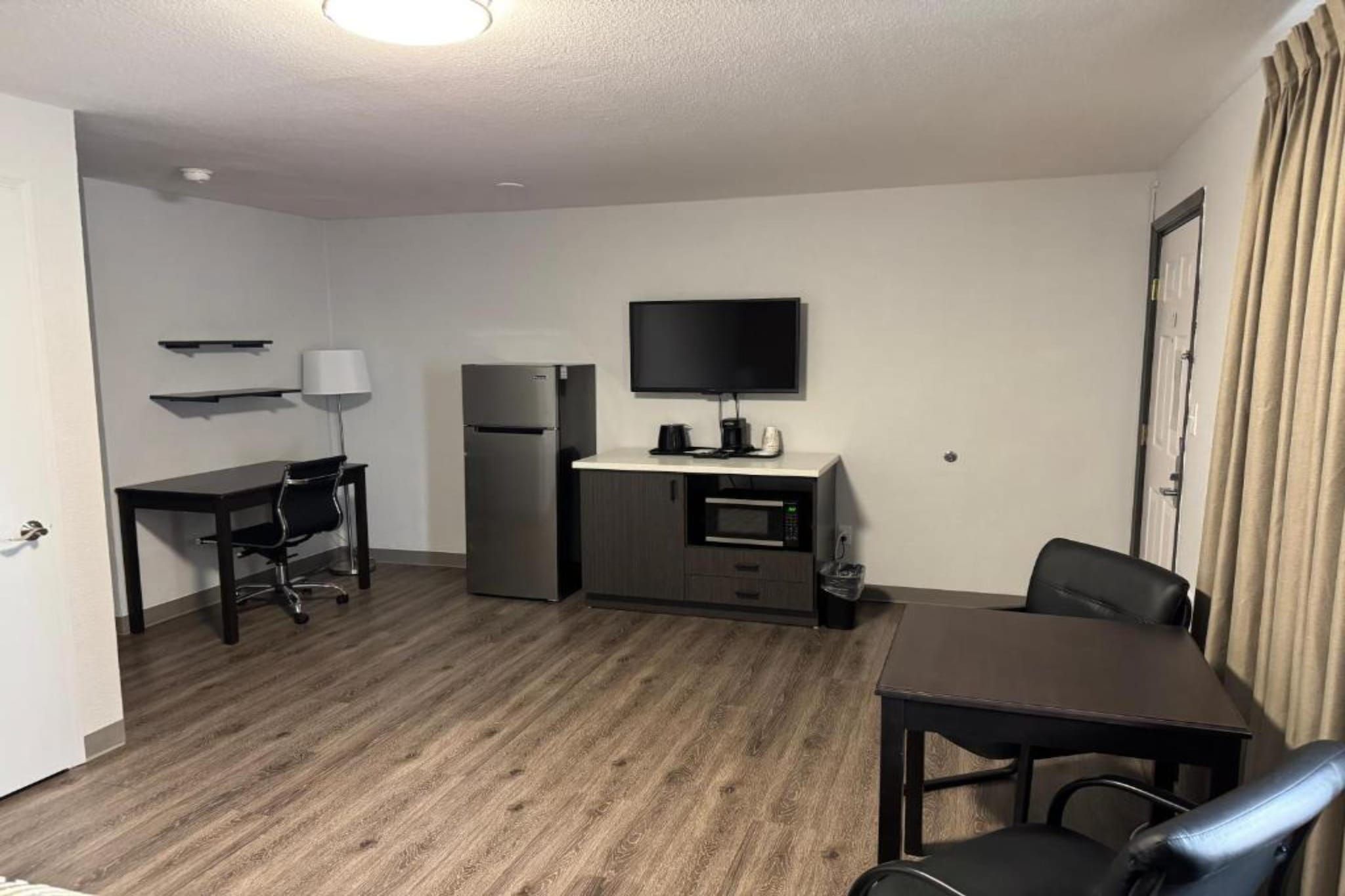 A compact hotel room with a kitchenette, a flat-screen TV, a desk, and a dining area.