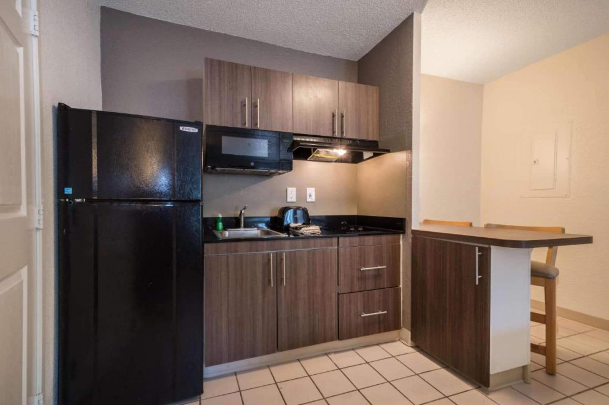 A compact kitchen with a black refrigerator, wooden cabinets, a microwave, and a small bar area.