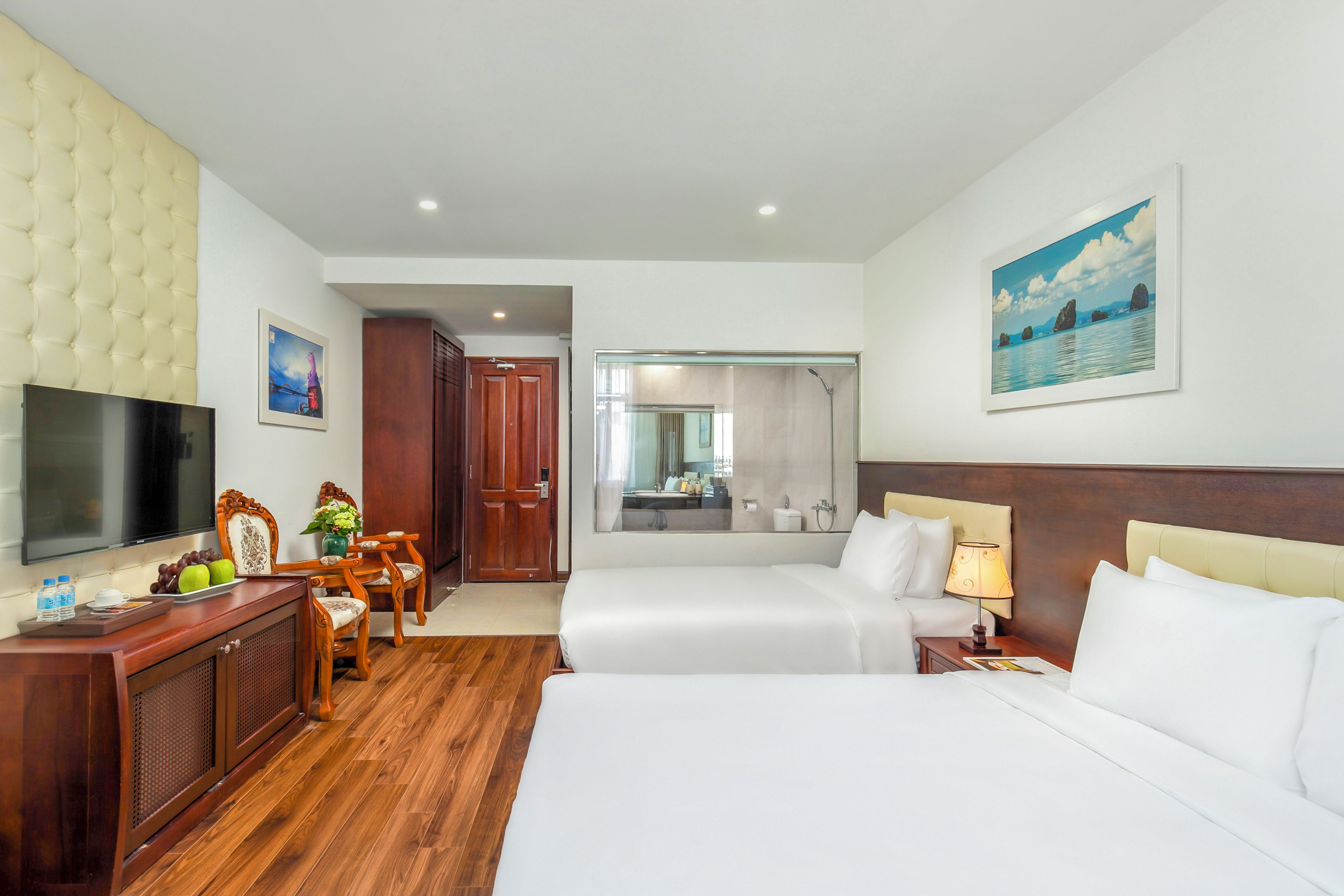 A hotel room with a bed, a desk with a TV, a bathroom visible through a glass door, and a painting on the wall.