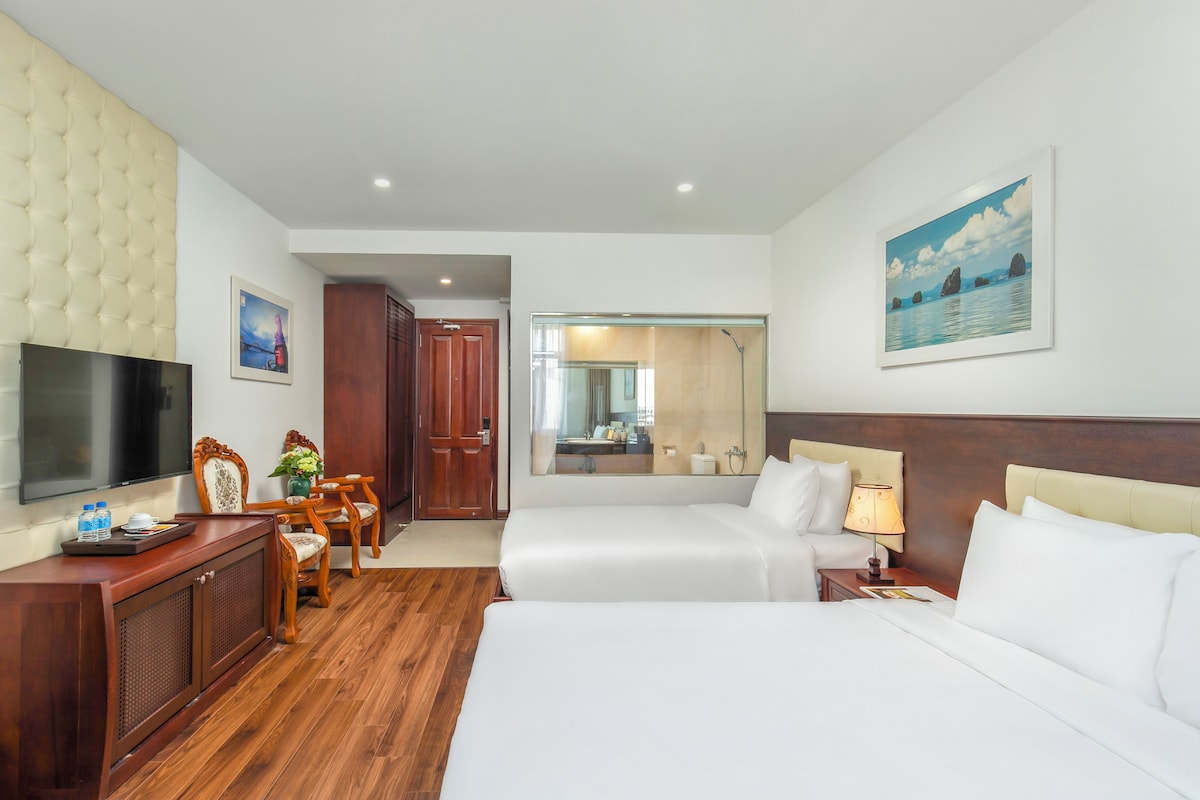 Une chambre d’hôtel comprenant un lit, un bureau avec une télévision, une chaise, une armoire en bois et une salle de bain visible à travers une porte en verre.