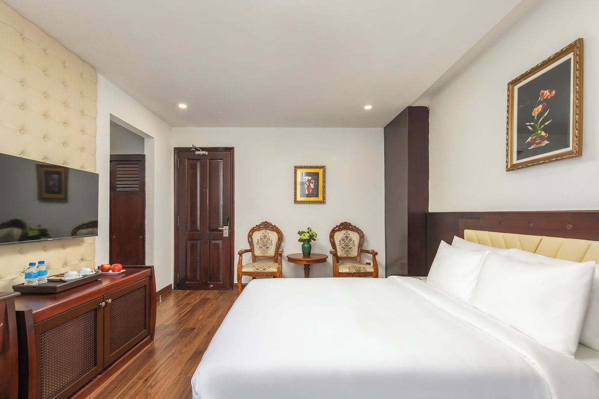 Une chambre d’hôtel avec un lit, deux chaises, une petite table avec un vase, une télévision et une porte.