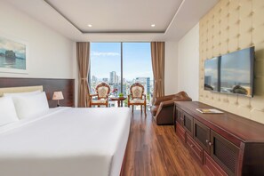 A hotel room with a large bed, a TV, a sofa, a dining table with chairs, and a city view. - Le Charmant Da Nang (An Hai)