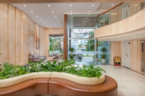 A spacious lobby with a curved seating area, a glass elevator, and a view of the cityscape through the glass. - Le Charmant Da Nang (An Hai)
