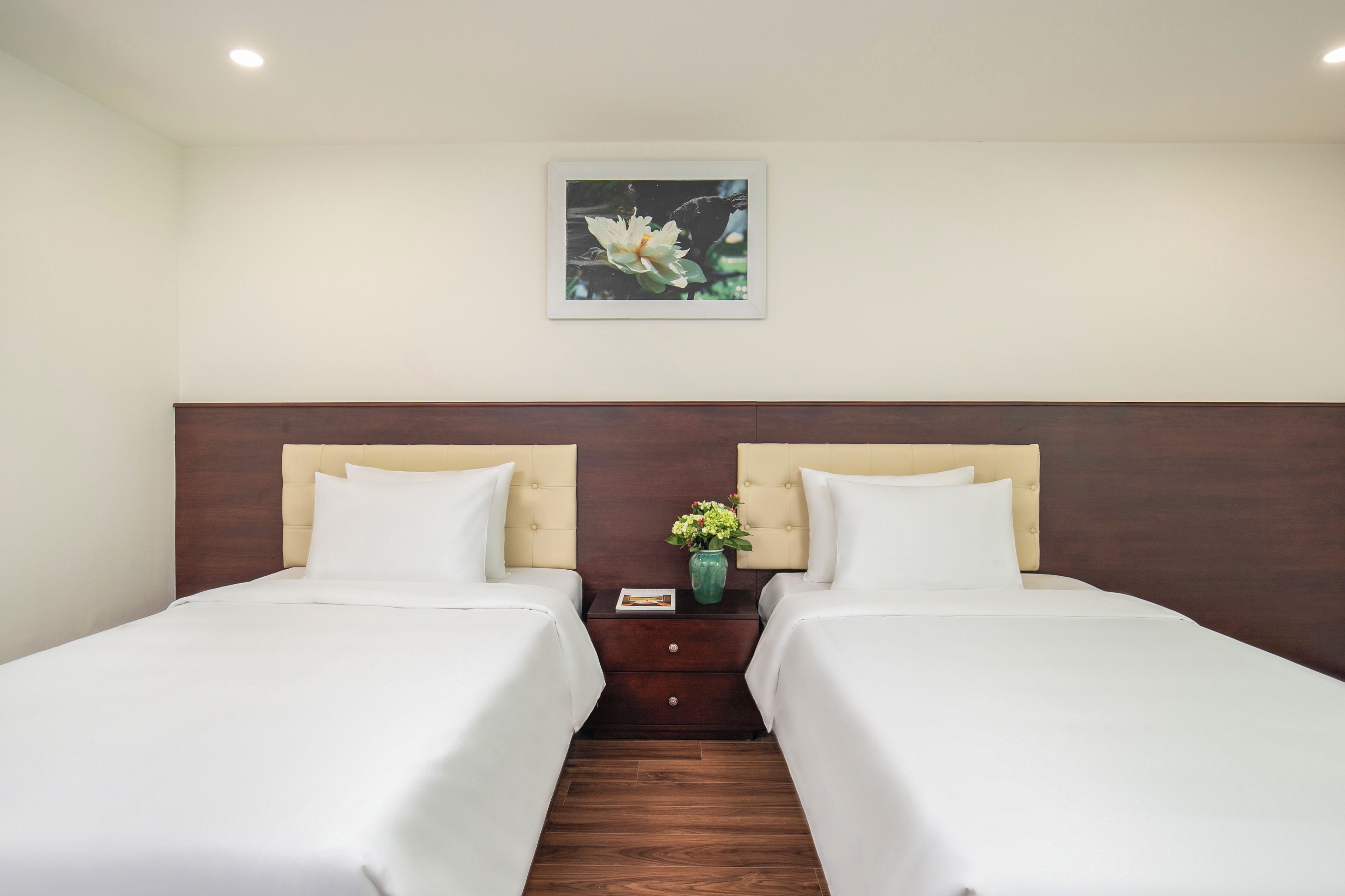 Quarto de hotel com duas camas, uma mesa de cabeceira com um vaso de flores e um quadro pendurado na parede.