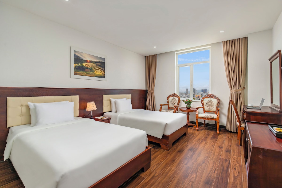 Une chambre d’hôtel avec deux lits, un bureau, une chaise, une fenêtre donnant sur la ville et un tableau au mur.