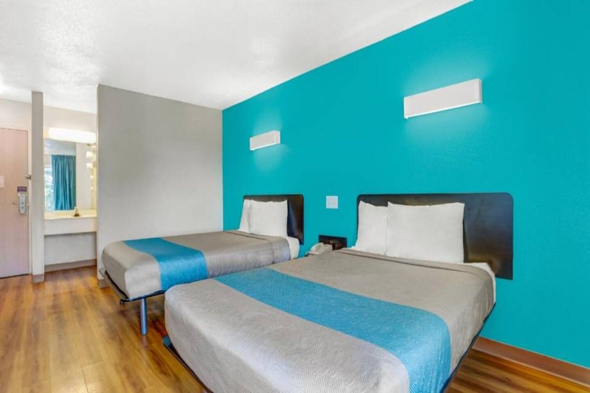 A hotel room with two beds, a wooden floor, a window with curtains, and a teal accent wall.