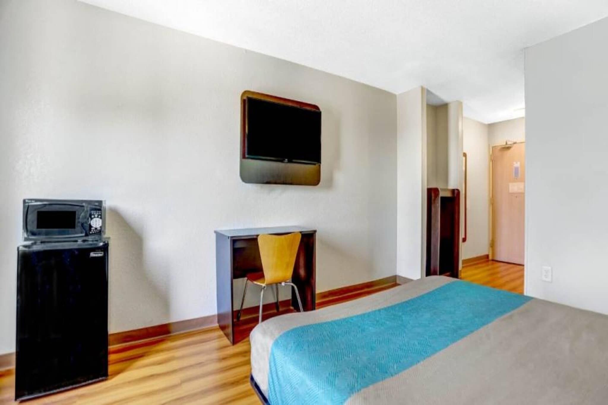 A hotel room with a bed, a desk, a chair, a television mounted on the wall, and a small refrigerator.