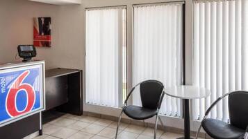A hotel room with a reception desk, a digital payment terminal, two chairs, a small table, and large windows with blinds.