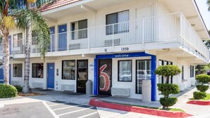 Um motel de dois andares com fachada azul e branca, numerado 1350, exibindo um grande número '6', uma varanda e um estacionamento.
