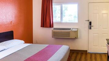 A hotel room with a bed, an air conditioning unit, a window with curtains, and a door.