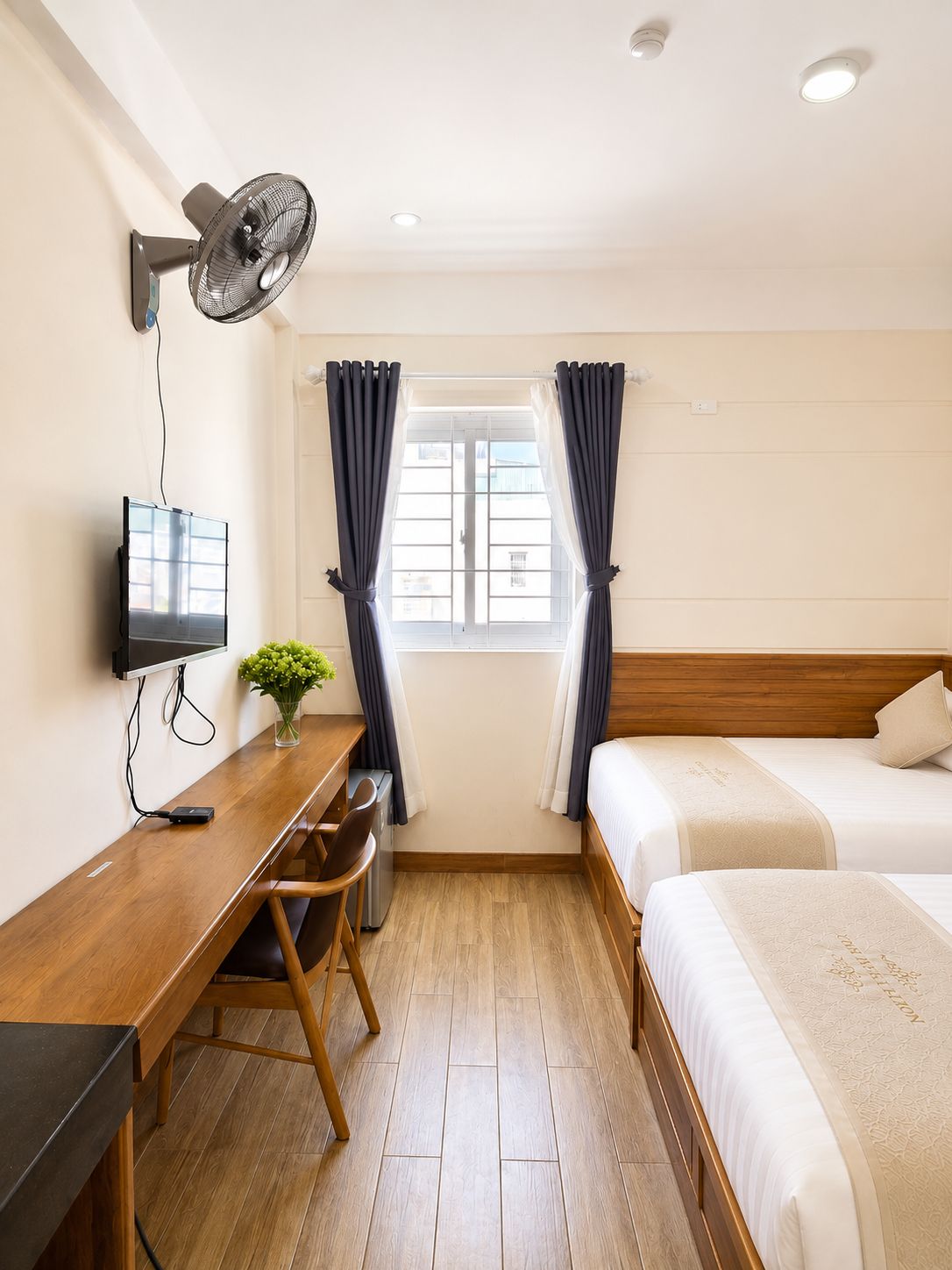A hotel room with a wooden desk, a bed, a TV, a wall-mounted fan, and a window with curtains.