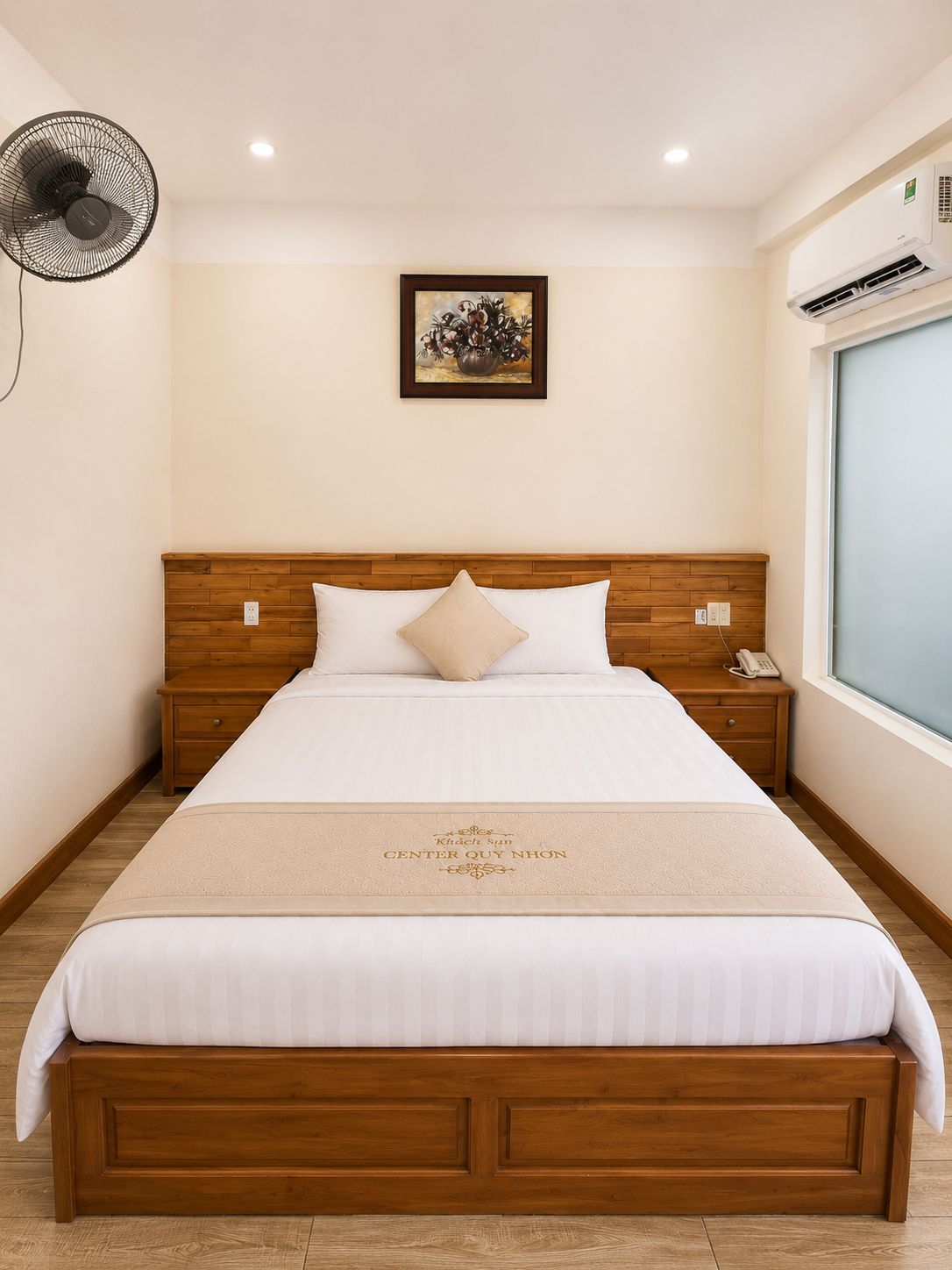 A neatly made bed with a wooden headboard, a bedside table with a phone, a wall-mounted fan, and a framed picture above the bed.